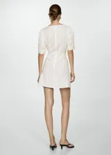 Vestido corto de color crudo con escote en pico y mangas cortas. Presenta un diseño de tejido con textura de cuadros y detalles bordados calados en el escote, las mangas y el ruedo. Cuenta con un corte recto y cierre de cremallera invisible en la parte posterior.