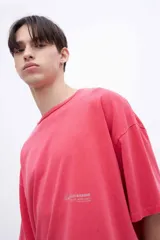 Remera de algodón teñida en prenda, color coral/rosado, con calce boxy y estampado en la espalda con texto en tipografía cursiva y líneas de texto más pequeñas.