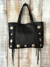 Cartera estilo tote color negro, confeccionada en cuero con textura granulada. Presenta detalles de tachas metálicas circulares en los laterales y el frente, junto con flecos largos de cuero que cuelgan de los costados. Cuenta con doble asa de hombro.