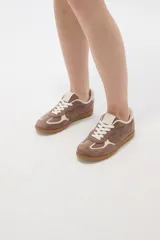 Zapatilla urbana de gamuza color marrón con detalles en beige, cordones claros y suela de goma tipo caramelo.