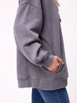 Canguro oversize de color gris con efecto lavado, capucha con cordones ajustables, bolsillo frontal tipo canguro y parche pequeño con logo de la marca en el pecho.