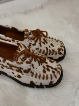 Zapato tipo mocasín de cuero con pelo estampado con diseño de manchas de vaca en tonos marrón y blanco. Presenta cordones frontales, costuras visibles en el borde y suela plana.