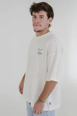 Remera color crema de corte holgado con cuello redondo y mangas cortas. Estampado en el pecho con la inscripción "Pulau Cold Winter" en letras azules y amarillas.