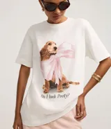 Remera blanca de algodón con cuello redondo y mangas cortas. Presenta una gran estampa central de un perro de raza cocker spaniel con un gran lazo rosa atado al cuello, y el texto "Do I look Pretty?" debajo.