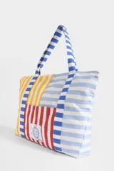 Bolso tipo tote de gran capacidad, confeccionado en algodón con diseño patchwork de rayas verticales y horizontales en colores amarillo, rojo, azul y blanco. Incluye un bolsillo frontal con un parche bordado con el escudo nacional.