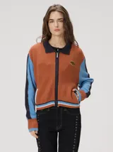 Campera estilo bomber Mutma Satifo, de diseño colorblock con cuerpo en color óxido y mangas celestes con detalles en azul marino. Cuenta con cuello camisero, cierre frontal, puños y ruedo elásticos rayados, y un parche gráfico en el pecho.