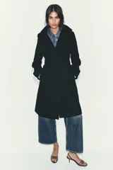 Abrigo de paño negro con capucha, cierre frontal cruzado con botones y cinturón de lazada en el mismo tejido. Presenta manga larga con trabillas en los puños y bolsillos laterales.