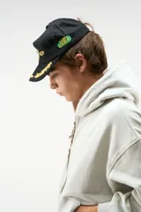 Gorra con visera plana y cierre snapback, confeccionada en algodón negro con corona no estructurada. Cuenta con detalles bordados en tonos dorados y verdes en el frente y la visera.