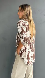 Blusa de corte holgado con estampado floral en tonos marrón y blanco. Presenta cuello camisero, cierre frontal con botones y un bolsillo en el pecho con detalle de lentejuelas. Las mangas son tres cuartos con trabas para ajustar.