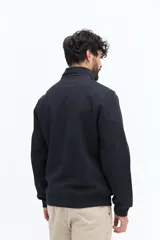 Campera negra de algodón con cierre frontal completo, cuello alto y bolsillos laterales con botón a presión. Presenta un pequeño parche rectangular con texto en el pecho izquierdo.