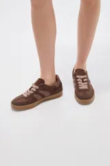 Zapatillas urbanas de corte bajo en tonos beige y tostado, combinando paneles de cuerina y gamuza sintética. Cuentan con cordones anchos tipo cinta de raso en color crudo, pespuntes decorativos y suela de goma al tono.