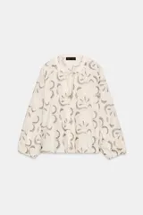 Blusa color crudo con bordados florales en azul oscuro. Presenta cuello redondo con escote pico ajustable mediante cordones con terminaciones de cuentas de madera, y mangas largas abullonadas con puños elásticos.