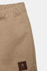 Pantalón de felpa color beige, tiro medio, con cintura elástica, bolsillos laterales ocultos y pierna ancha. Presenta un parche pequeño con un logo en la parte delantera.