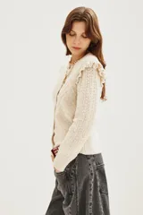 Cardigan de punto color beige con textura calada, cuello redondo y detalle de volados en los hombros. Presenta cierre frontal y puños acanalados.
