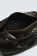 Bolso de viaje de diseño rectangular con acabado craquelado en tono oscuro. Cuenta con doble asa de mano, compartimento principal con cierre de cremallera, bolsillo interior con cierre y correa de hombro ajustable y extraíble.