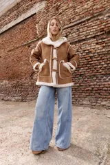 Campera estilo gamulán color tostado con interior y bordes de corderito sintético color crema. Presenta capucha amplia, cierre frontal con trabas y broches metálicos, y bolsillos delanteros grandes. Posee un calce relajado y diseño de textura suave.