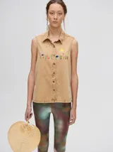 Camisa sin mangas tipo musculosa confeccionada en denim liviano de color tostado. Presenta cuello camisero y cierre frontal con botones. Destaca por sus bordados coloridos en el pecho con motivos veraniegos como sol, olas y elementos marinos.