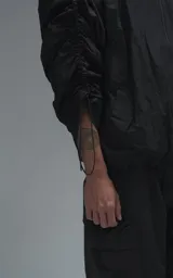 Campera rompevientos negra de calce oversize, confeccionada en microfibra técnica ligera con acabado semi-mate. Presenta capucha integrada, cierre frontal técnico y bolsillos tipo ojal. Destaca por su sistema de frunces elásticos regulables mediante tancas a lo largo de las mangas y el ruedo, permitiendo ajustar el volumen de la prenda.