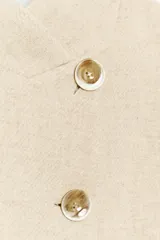 Blazer de corte estructurado en color beige, con cuello mao y cierre frontal cruzado mediante botones. Presenta mangas largas con detalle de botones en los puños y bolsillos de vivo en el frente.