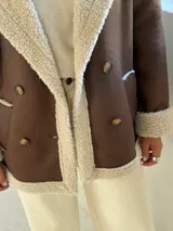 Tapado de estilo aviador en color chocolate con interior y detalles de corderito en color beige. Presenta cierre frontal cruzado con botones, solapas amplias y puños con borde de corderito.