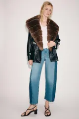 Trench corto Zara de efecto piel color negro, con cuello de solapa y aplicación de pelo sintético a contraste en color marrón. Cuenta con manga larga con trabillas en los puños, bolsillos delanteros y cinturón ajustable del mismo tejido.