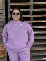 Canguro de tela deportiva color lavanda, con capucha, cordones ajustables en contraste y logo bordado en el pecho.