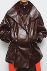 Chaqueta amplia de efecto piel en color marrón oscuro, con cuello alto, manga larga y bolsillos delanteros. Presenta cintura ajustable, forro interior y cierre frontal con cremallera y botones ocultos por solapa.