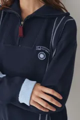 Buzo de tejido de punto color marrón con cuello alto y cierre frontal parcial. Presenta rayas verticales finas en color claro, puños y ruedo en tejido acanalado color crema, y detalles en color verde lima en el tirador del cierre, el cordón de ajuste del ruedo y el parche bordado en el pecho.