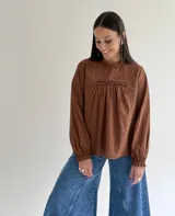 Blusa de color marrón con cuello redondo, mangas largas con puños elásticos y detalle de puntillas verticales en el frente.