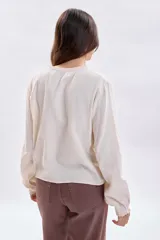 Blusa de estilo romántico en color crudo, confeccionada en tela texturizada con detalles de encaje y alforzas en el canesú. Presenta cuello redondo, cierre frontal con botones y mangas largas con puños delicados.