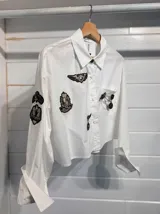 Camisa blanca de algodón con corte cropped y calce relajado. Presenta cuello clásico, mangas largas con puños anchos y una serie de parches bordados estilo militar e insignias metálicas decorativas en el frente y las mangas.