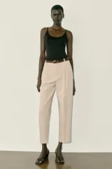 Pantalón tobillero de tiro medio, color beige, con pinzas delanteras y bolsillos frontales. Incluye un cinturón fino de cuero marrón con hebilla metálica dorada y cierre frontal con cremallera, botón interior y gancho.