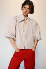 Blusa de cuello subido con escote en pico y cierre frontal con botones forrados. Presenta mangas abullonadas de largo tres cuartos con elástico en los puños y detalle de lazos en el cuello y el frente.