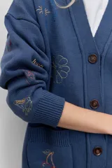 Cardigan de punto azul con cuello en V, cierre frontal de botones y bolsillos delanteros. Presenta bordados decorativos con motivos variados y frases en colores contrastantes.