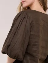 Blusa color beige confeccionada en lino, con escote en V pronunciado y cierre frontal de múltiples lazos para atar. Presenta mangas abullonadas tres cuartos con puños elásticos y corte relajado.