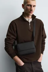 Bandolera verde militar con acabado engomado y textura geométrica. Tiene un compartimento principal con cierre de solapa y botón a presión, un bolsillo interior con cremallera y un asa de hombro ajustable con monedero.