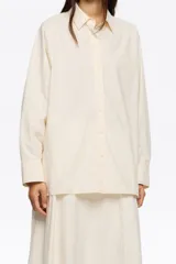 Camisa oversize de algodón color crema, con cuello camisero, botones frontales y mangas largas ranglán con puños abotonados. Presenta pespuntes a contraste en toda la prenda.