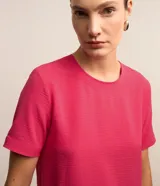 Blusa fucsia de manga corta con cuello redondo pespunteado, diseño sin estampa y efecto airflow con textura sutil. Cuenta con abertura trasera tipo gota y cierre con botón.