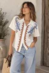 Camisa beige de manga corta con cuello y cierre frontal de botones. Presenta delicados bordados de hojas y flores en tonos verdes y naranjas a los lados, y una guarda ornamental ondulada alrededor de la botonera.