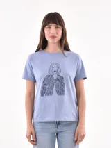 Remera azul claro de manga corta con estampado frontal de una mujer con cabello rubio y una chaqueta a rayas.