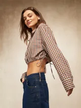 Camisa de manga larga, cuello camisero y botones frontales, con estampado a cuadros marrones y blancos. El ruedo inferior es ajustable con cordones laterales, creando un efecto *cropped* y fruncido.