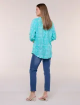 Blusa turquesa de silueta suelta confeccionada en tejido liviano, con escote en V, pequeña cartera frontal con botones decorativos y mangas 3/4 con presilla para arremangar. Presenta patrón estampado geométrico tipo tie-dye y ruedo curvo.