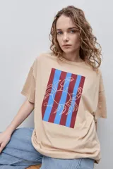 Remera color beige de silueta amplia y calce relajado, con cuello redondo y mangas cortas de hombros caídos. Presenta una estampa frontal rectangular con rayas verticales en tonos bordó y celeste, sobre la cual se superpone el dibujo lineal de un cangrejo.