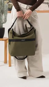 Cartera tipo hobo de tamaño grande en color beige, con detalles en negro. Presenta una franja horizontal negra con el logo de la marca, asa corta de hombro y correa larga desmontable. Incluye bolsillo frontal con cierre oculto y bolsillo trasero.