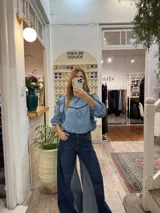 Blusa de jean fino con cuello alto, mangas largas con volumen y volados decorativos en los hombros. Presenta detalles de ribetes en terciopelo azul oscuro en el frente y los volados.