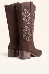 Bota de caña alta estilo tejano confeccionada en gamuza color marrón oscuro. Presenta detalles de costuras decorativas en forma de llama en la caña, taco cuadrado de madera y punta almendrada.