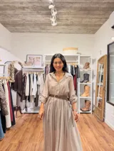 Vestido largo de seda en color beige, con mangas tres cuartos, cuello camisero y falda amplia con caída fluida. Incluye un cinturón tejido en tono marrón que marca la cintura.