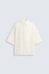 Camisa de corte relajado (relaxed fit) color crema o blanco roto, confeccionada en tejido con mezcla de algodón. Presenta un bordado sutil en relieve con patrón de ondas o líneas diagonales entrecruzadas. Tiene cuello solapa, manga corta y un bolsillo de plastrón en el pecho.
