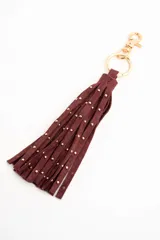 Charm para bolso compuesto por flecos de cuero sintético color marrón, decorados con pequeñas tachas metálicas doradas. Incluye un aro y un cierre de mosquetón metálico en tono dorado.