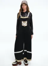 Mono largo de color negro con corte holgado y perneras anchas. Presenta detalles de encaje blanco y tela estampada floral en los tirantes, el bolsillo frontal, la parte inferior de las piernas y el ruedo.